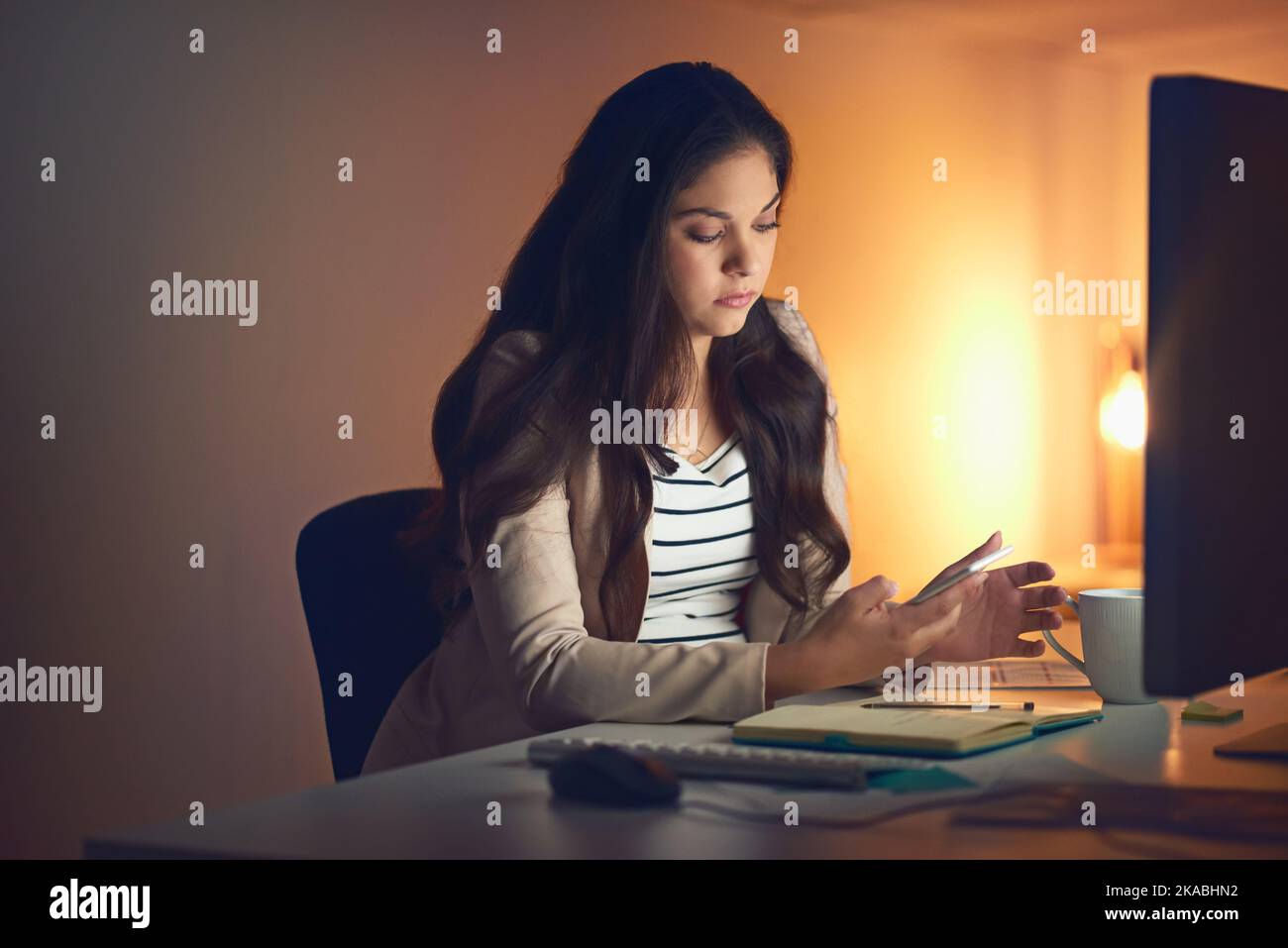 Wir müssen manchmal härter arbeiten. Eine junge Geschäftsfrau, die während einer späten Nacht bei der Arbeit ein Mobiltelefon und einen Computer benutzt. Stockfoto