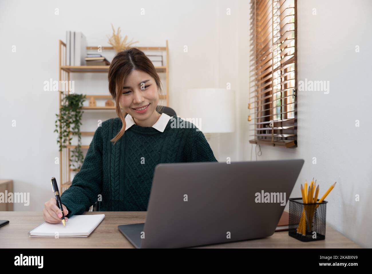 asiatische Studentin, die fernab von zu Hause studiert, einen Laptop benutzt, während des Online-Unterrichts Notizen auf dem Notizblock macht, E-Learning-Konzept, lächelnd Stockfoto