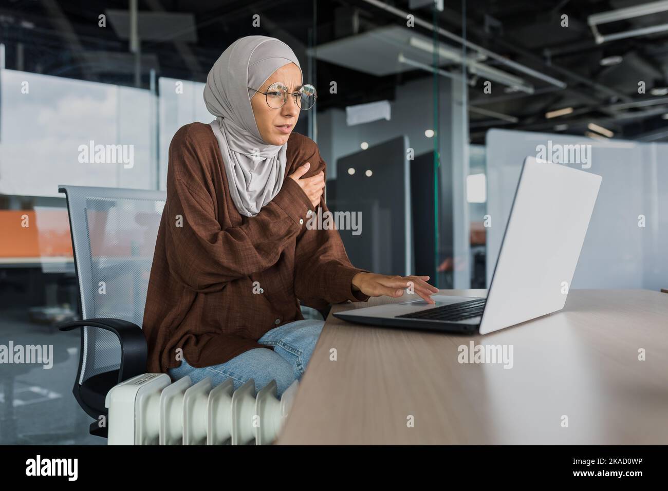 Geschäftsfrau friert bei der Arbeit im Büro ein, Frau in Hijab in der Nähe der Heizung, die versucht, warm zu arbeiten, im Bürogebäude mit Laptop, kalt im Büro. Stockfoto