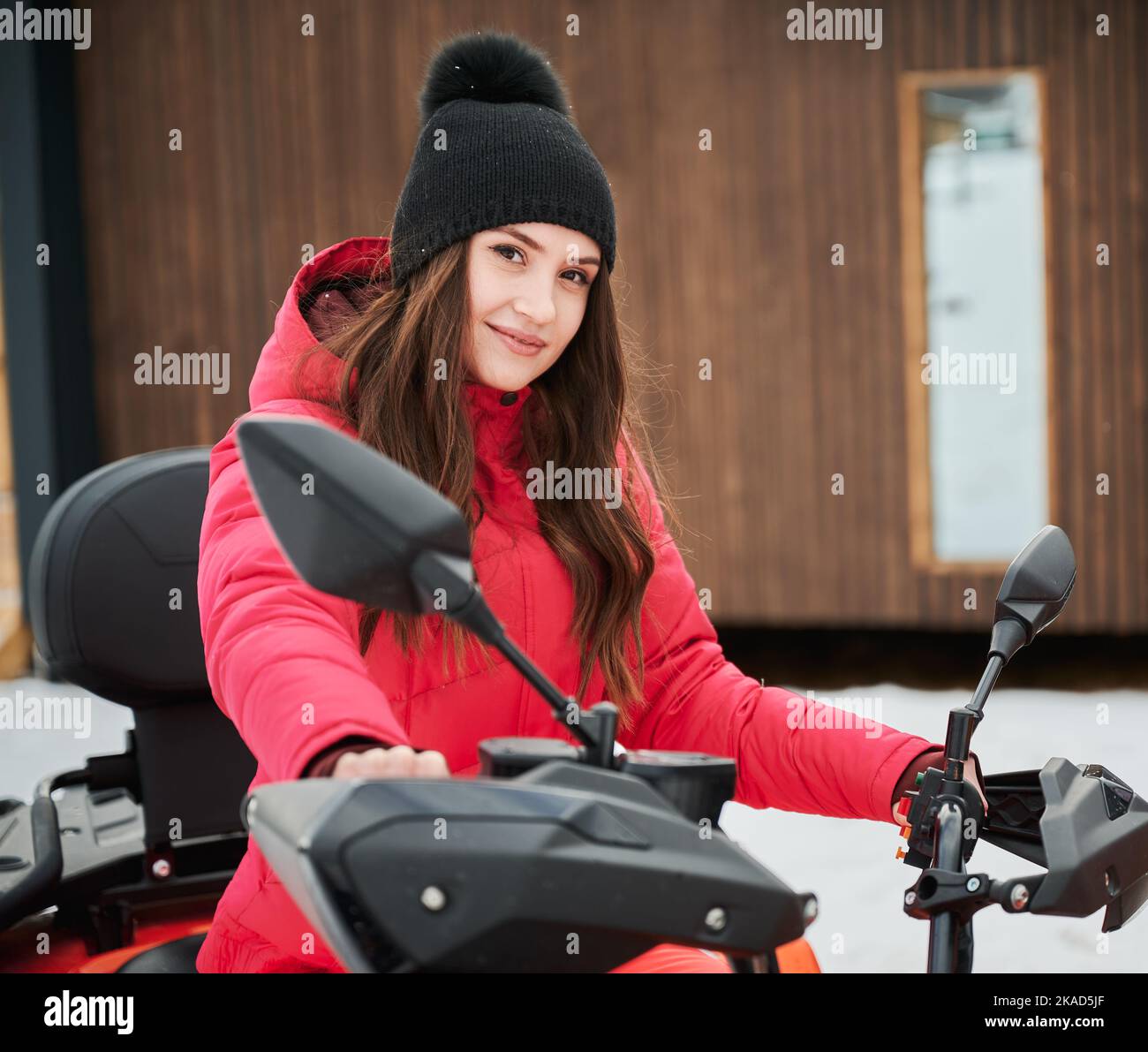 Porträt einer schönen Frau, die auf Geländewagen-Geländewagen-ATV mit Holzhaus im Hintergrund posiert. Konzept der aktiven Freizeit und Winteraktivitäten. Stockfoto