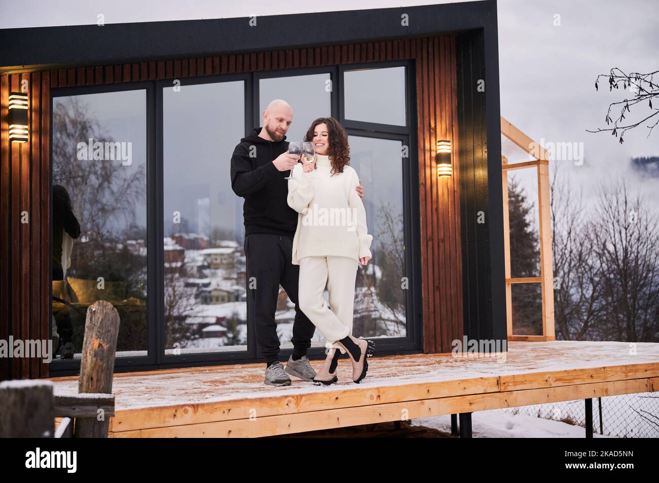 Ein Paar liebt es, vor dem Holzhaus mit Panoramafenster einen alkoholischen Drink zu genießen. Glückliche Frau und Mann klirren Gläser Wein und feiern gemeinsam am Wintertag in den Bergen. Stockfoto