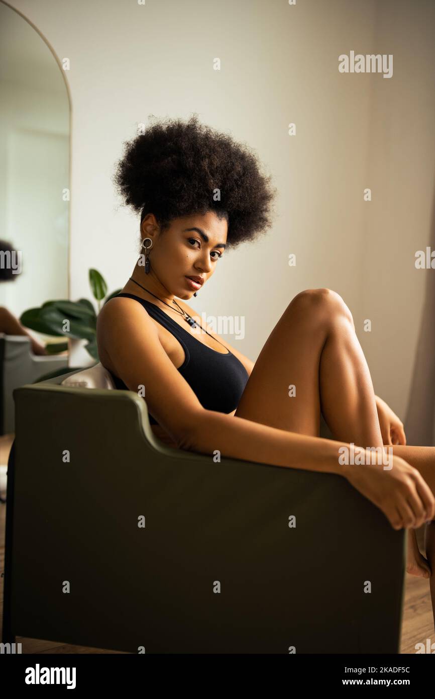 Lockige afrikanisch-amerikanische Frau in schwarzem Crop Top, die Kamera anschaut, während sie zu Hause auf einem Sessel sitzt, Stockbild Stockfoto