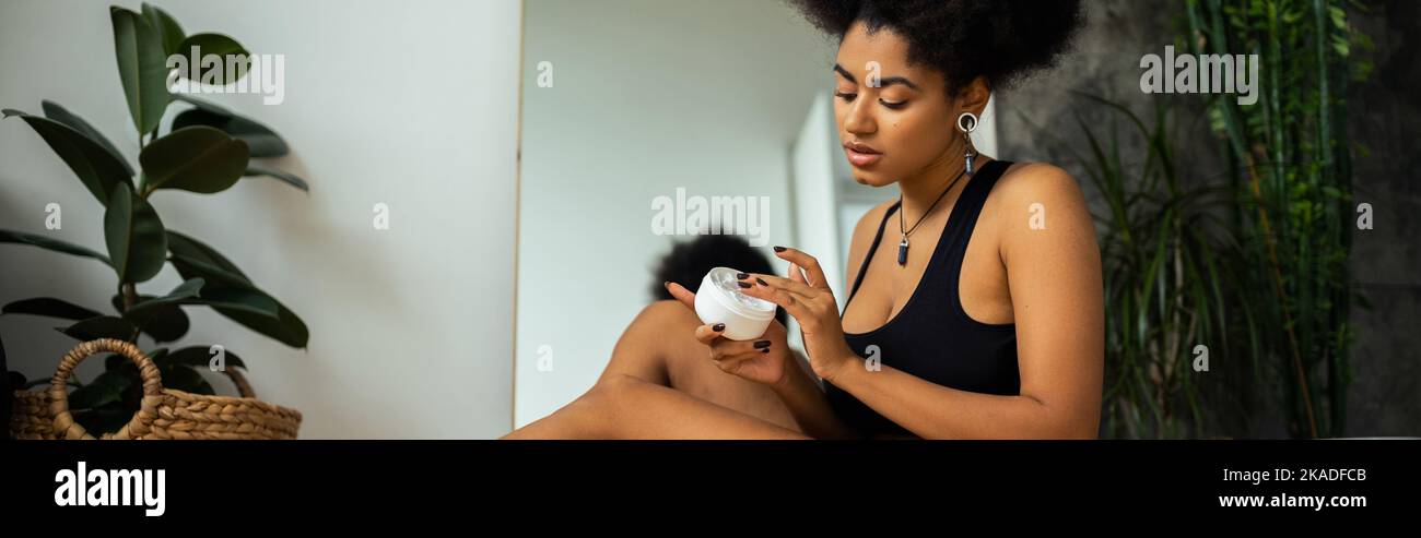 Lockige afroamerikanische Frau hält kosmetische Creme in der Nähe Spiegel und Pflanzen im Badezimmer, Banner, Stock Bild Stockfoto