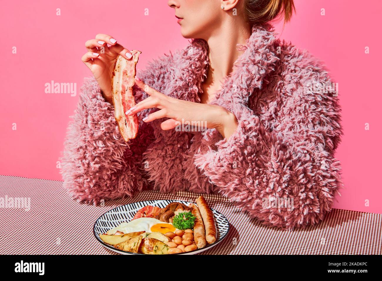 Stilvolle Frau in rosa pelzigen Mantel essen amerikanisches Frühstück mit Spiegelei, Würstchen und Speck auf rosa Hintergrund Stockfoto