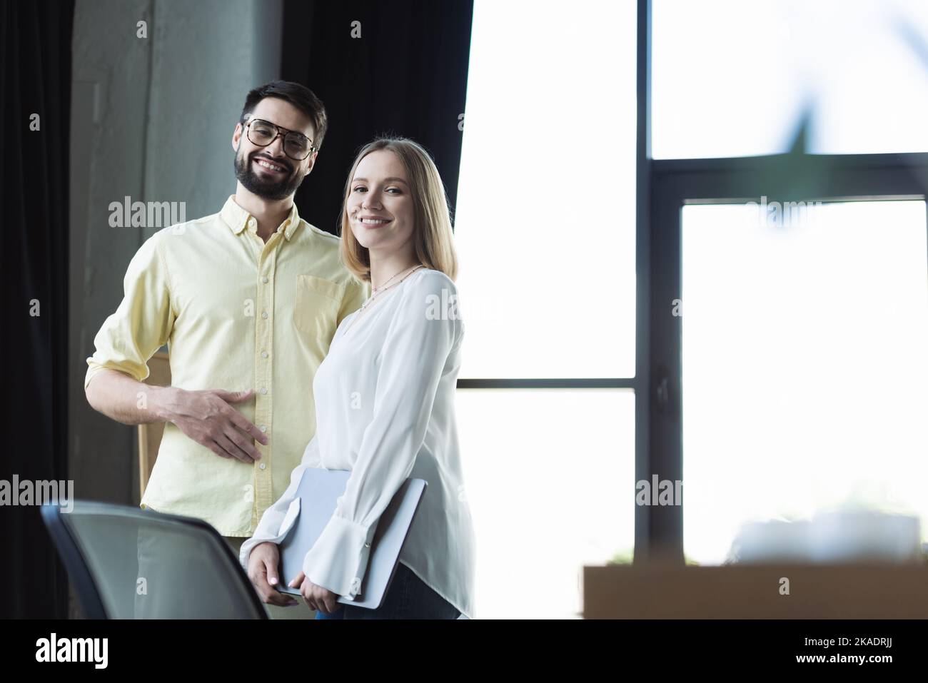 Fröhlicher Geschäftsmann, der die Kamera in der Nähe eines neuen Mitarbeiters mit einem Laptop im Büro anschaut, Bild aus dem Stock Stockfoto