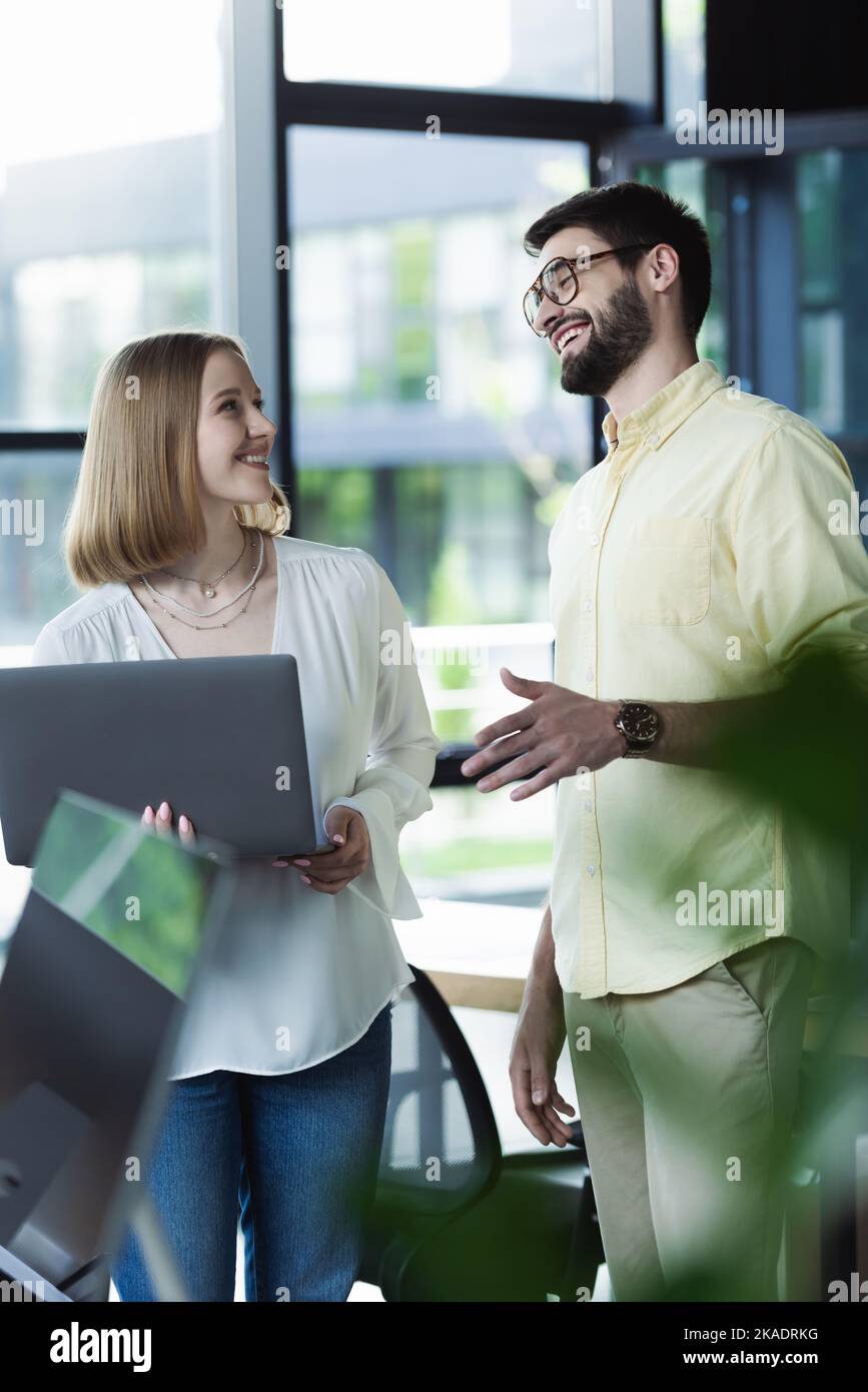 Fröhlicher Geschäftsmann im Gespräch mit Praktikant mit Laptop im Büro, Bild aus dem Stock Stockfoto