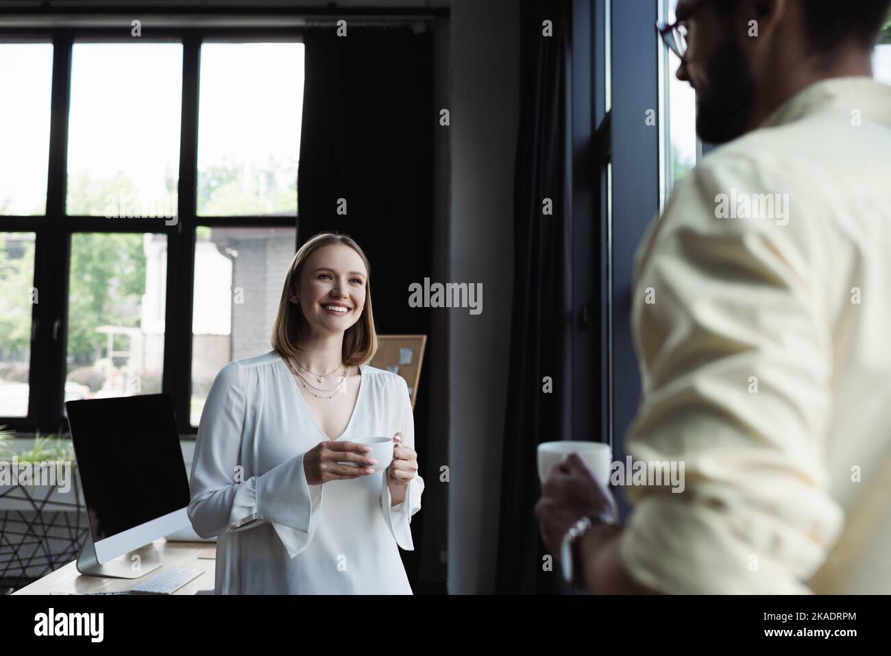 Fröhlicher neuer Arbeiter, der eine Tasse Kaffee in der Nähe eines verschwommenen Geschäftsmannes im Büro hält, Stockbild Stockfoto