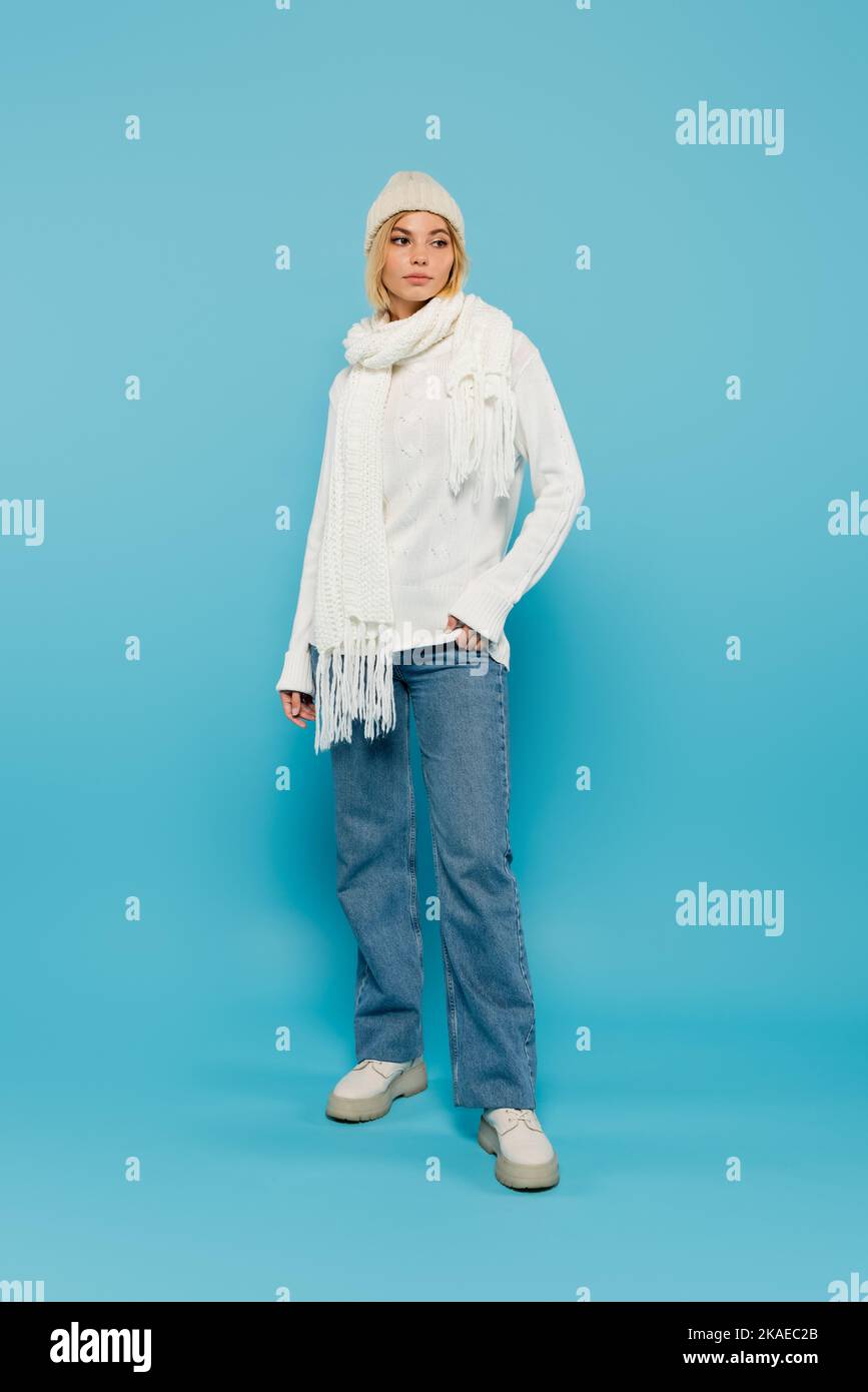 Volle Länge der jungen blonden Frau in weißen Pullover und Winterhut stehen mit Hand in der Tasche auf blau, Stock Bild Stockfoto