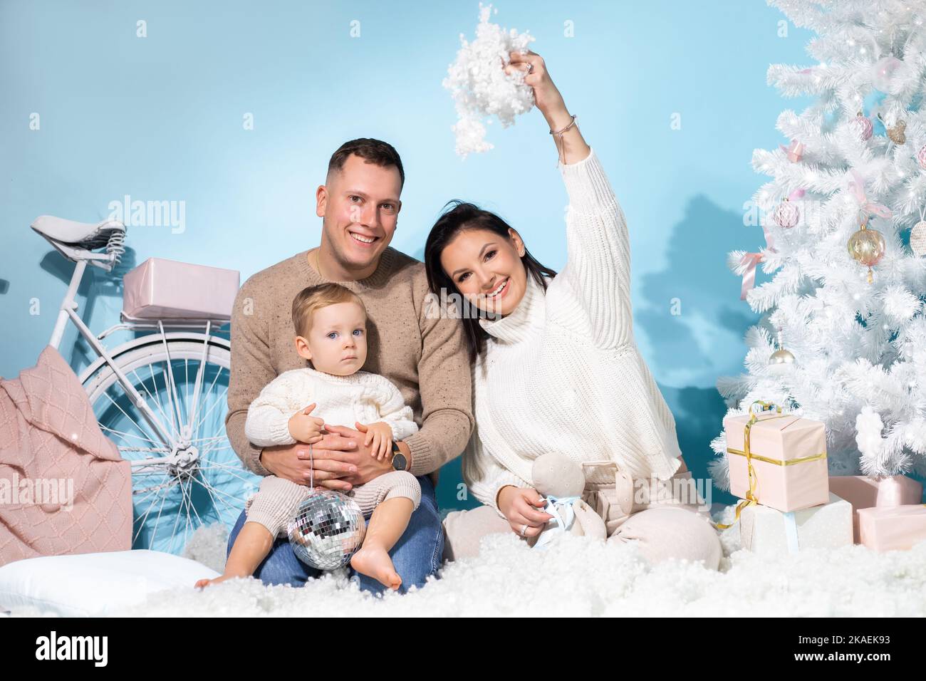 Familienportrait von drei Personen, die zusammen in einem weihnachtlichen Drehort mit weißen Dekorationen auf blauem Hintergrund sitzen. Fröhliche Mutter, Vater und Stockfoto