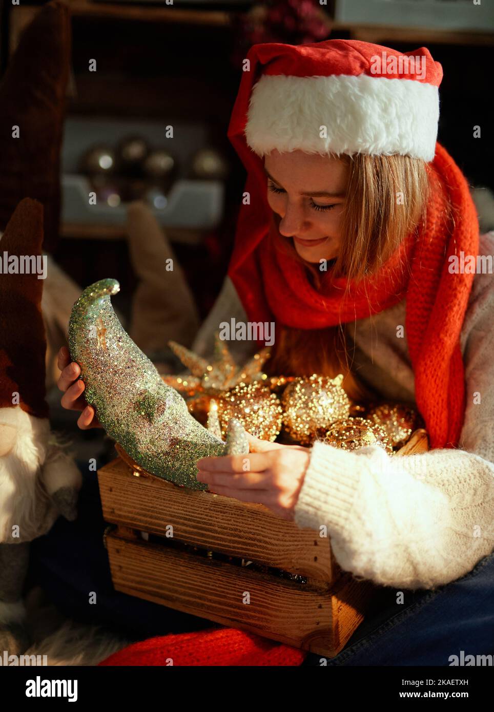 Fröhlicher, fröhlicher kaukasischer Teenager in Weihnachtsmütze und rotem Schal mit Neujahrsdekoration in Holzkiste und schwachem Licht. Mädchen Auspacken Winter Weihnachtsdekoration zu Hause in der Nähe Weihnachtsbaum. Hochwertige Fotos Stockfoto