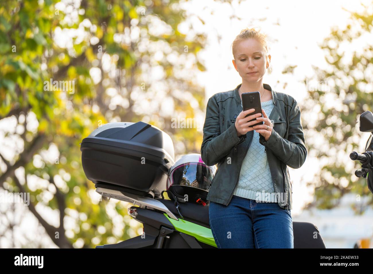 Rothaarige Frau, die das Smartphone neben ihrem geparkten Motorrad benutzt Stockfoto