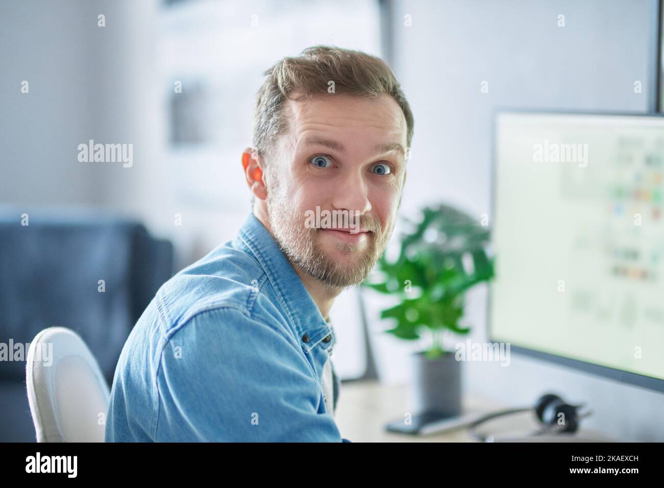 Niedlicher bärtiger Kaukasier, freiberuflicher Software-Ingenieur, der am Arbeitsplatz im Heimbüro sitzt und kaum mit dem Computer arbeitet. Junger, hübscher Entwickler-Mann im Denim-Hemd. Hochwertige Bilder Stockfoto