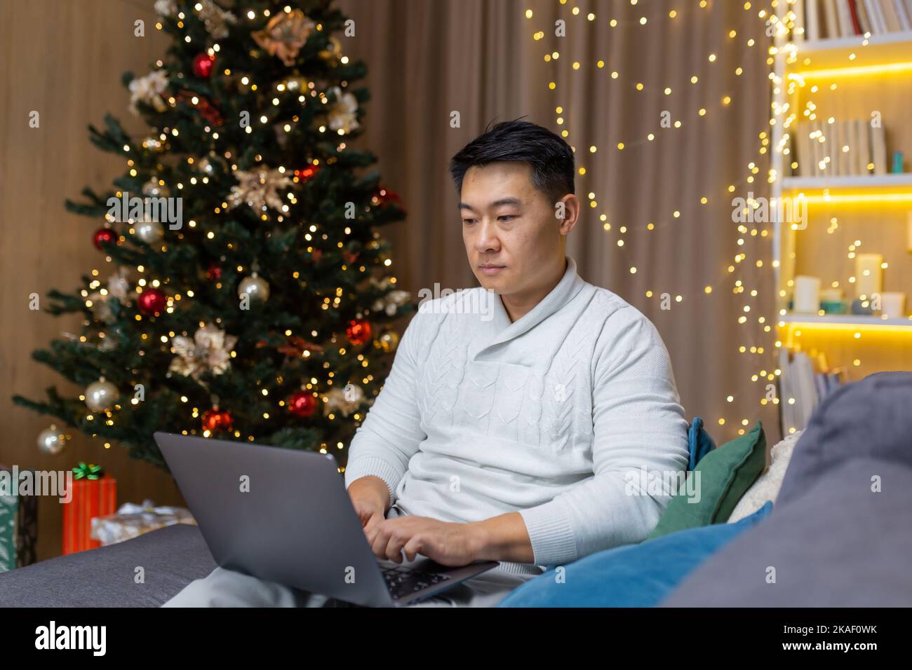 Ein junger asiatischer Geschäftsmann arbeitet im Urlaub an einem Laptop. Er sitzt ernst zu Hause auf dem Sofa in der Nähe des Weihnachtsbaums. Stockfoto