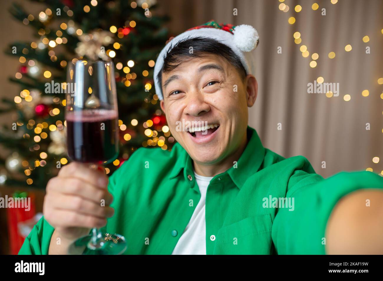Glücklicher junger, gutaussehender asiatischer Mann in rotem Weihnachtsmannhut und grünem Hemd, der zu Hause in der Nähe des Weihnachtsbaums Selfie macht, ein Glas Wein hält und lächelt. Stockfoto