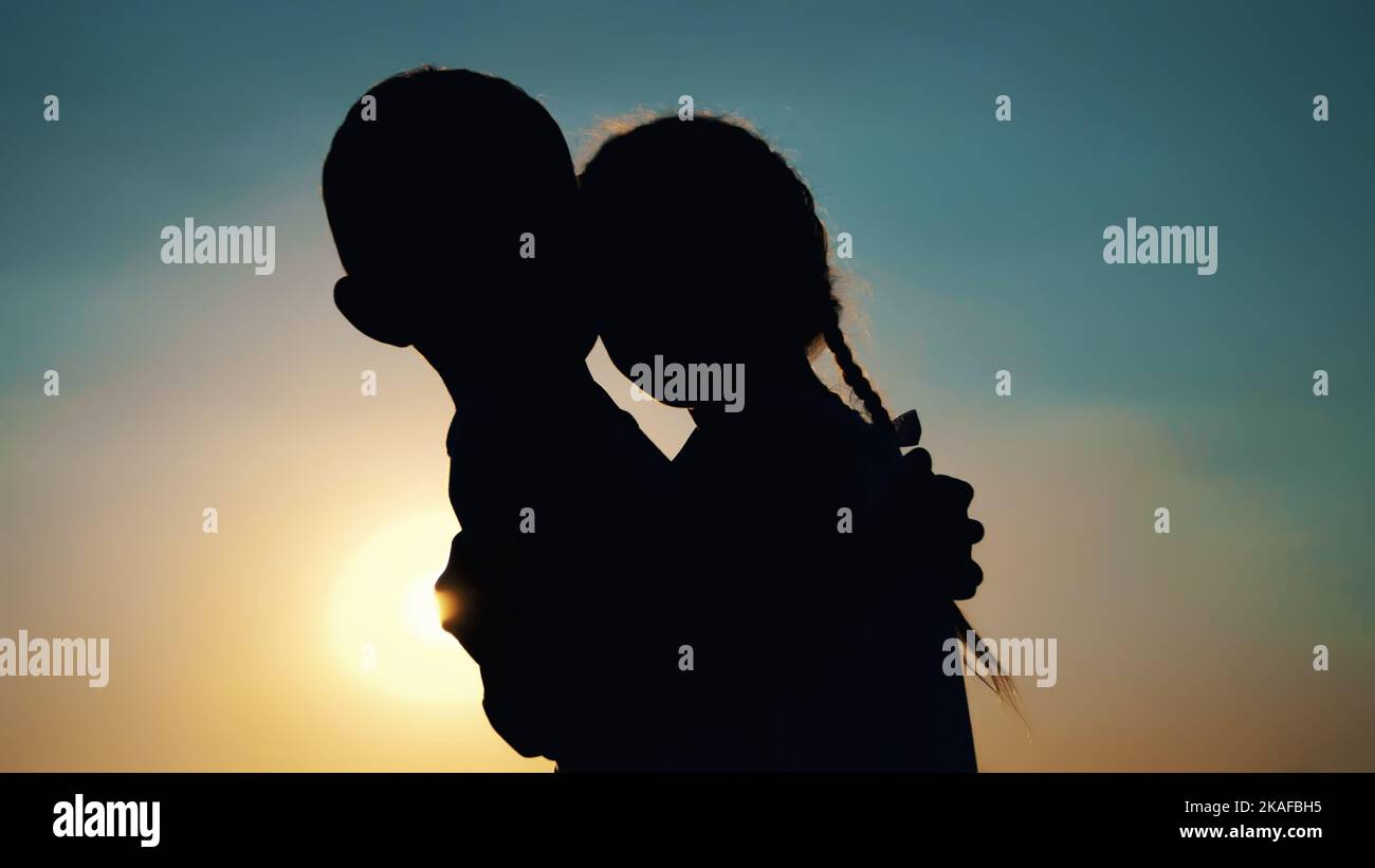 Silhouetten, Figuren von Kindern, Jungen und Mädchen, Bruder und Schwester, die sich vor dem Hintergrund der Sonne umarmen, bei Sonnenuntergang, im Sommer. Hochwertige Fotos Stockfoto