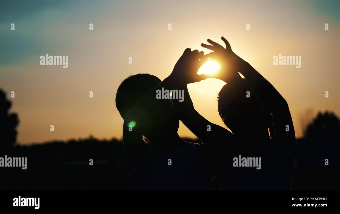 Silhouetten, Figuren von Kindern, Jungen und Mädchen zeigen die Herzen mit den Fingern, Händen, vor dem Hintergrund der Sonne, bei Sonnenuntergang im Sommer. Frohe Familie. Zeitlupe. Hochwertige Fotos Stockfoto