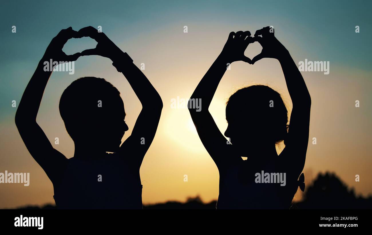 Silhouetten, Figuren von Kindern, Jungen und Mädchen zeigen die Herzen mit den Fingern, Händen, vor dem Hintergrund der Sonne, bei Sonnenuntergang im Sommer. Frohe Familie. Zeitlupe. Hochwertige Fotos Stockfoto