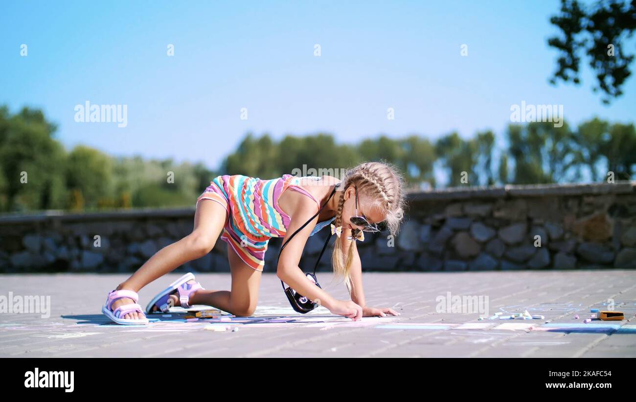 Ein Mädchen in Sonnenbrillen, zeichnet Zeichnungen mit farbigen Buntstiften auf den Asphalt, Straßenfliesen. Ein heißer Sommertag. Hochwertige Fotos Stockfoto