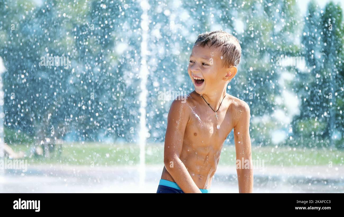 Aufgeregt Junge von sieben Jahren Spaß zwischen Wasserstrahlen, in Brunnen, herumlaufen, streuen, Spaß haben, Viel Spaß an einem heißen Sommertag. Sommer in der Stadt. Zeitlupe. Hochwertige Fotos Stockfoto