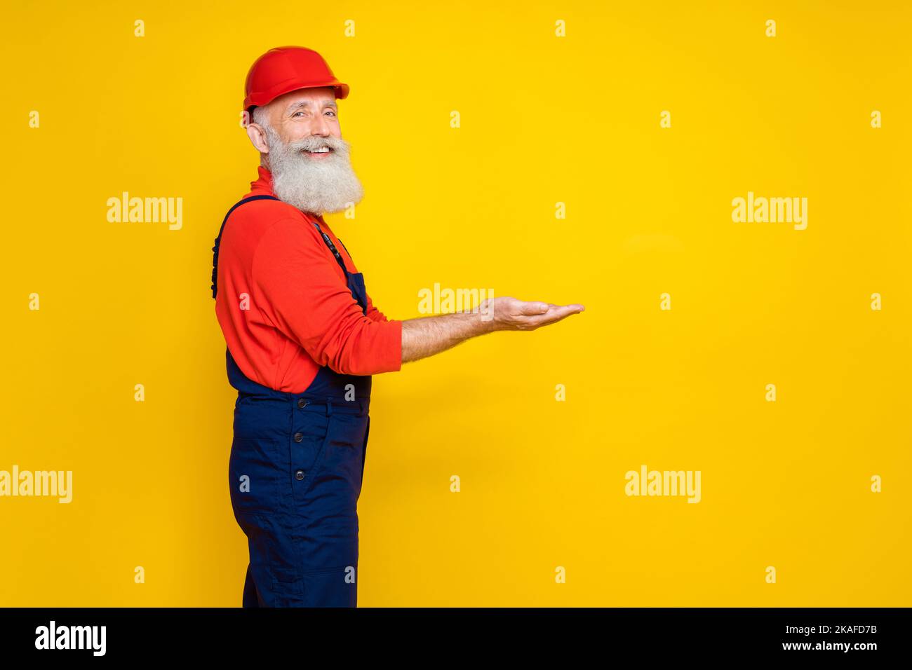 Foto von schönen niedlichen Alter Mann Arbeitskleidung insgesamt roten Hut hält Arme leeren Raum isoliert gelbe Farbe Hintergrund Stockfoto