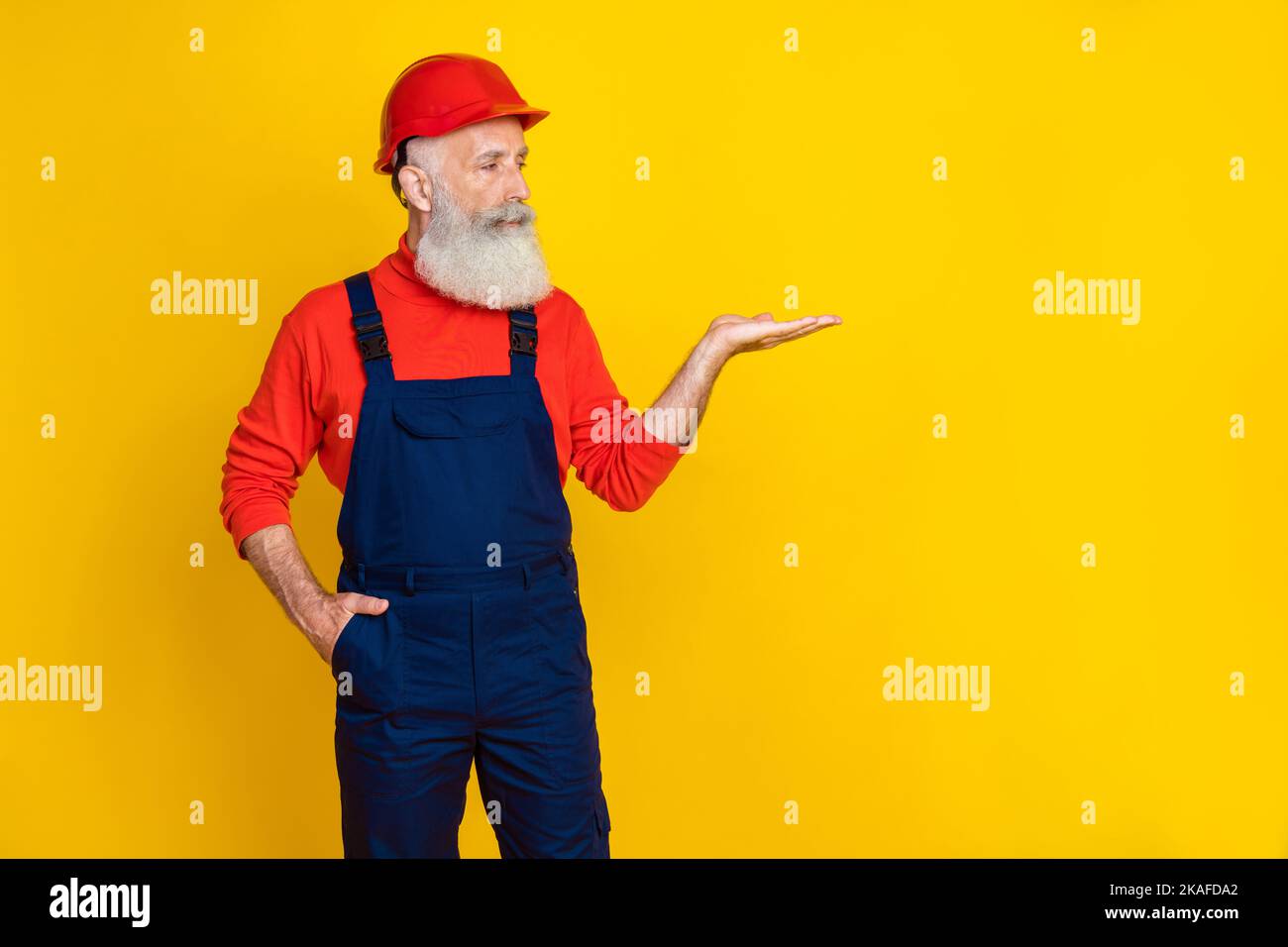 Foto von fröhlich positive Senior guy gekleidet Uniform insgesamt rot hardhat suchen Arm leeren Raum isoliert gelben Hintergrund Stockfoto