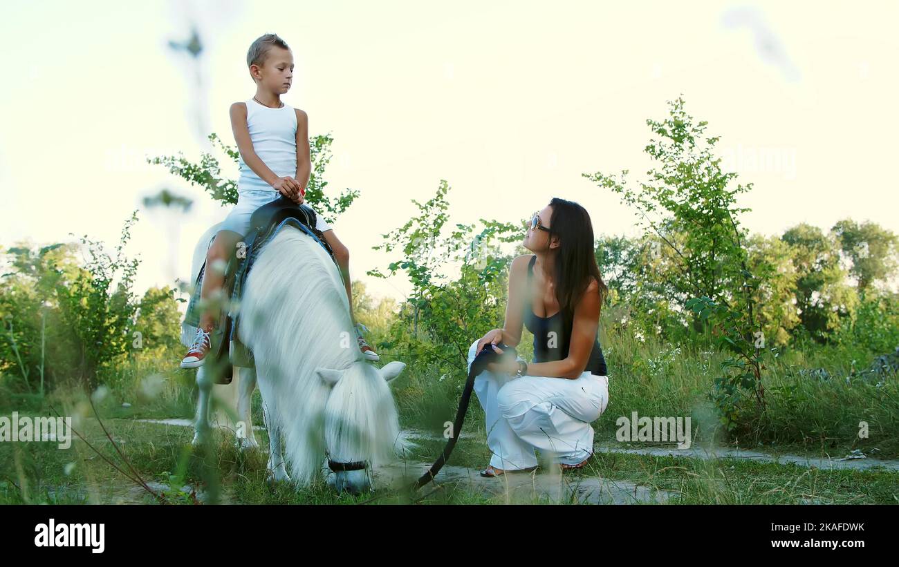 Eine Frau und ein Junge gehen um das Feld, Sohn reitet auf einem Pony, Mutter hält ein Pony für einen Zaum. Fröhlicher, glücklicher Familienurlaub. Im Freien, im Sommer, in der Nähe des Waldes. Hochwertige Fotos Stockfoto