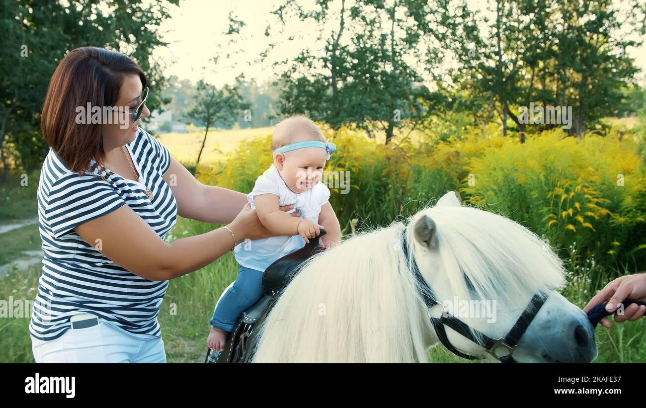 Eine Frau mit einem Baby geht um das Feld, ein Baby sitzt auf einem Pony, Mama hält das Baby. Fröhlicher, glücklicher Familienurlaub. Im Freien, im Sommer, in der Nähe des Waldes. Hochwertige Fotos Stockfoto