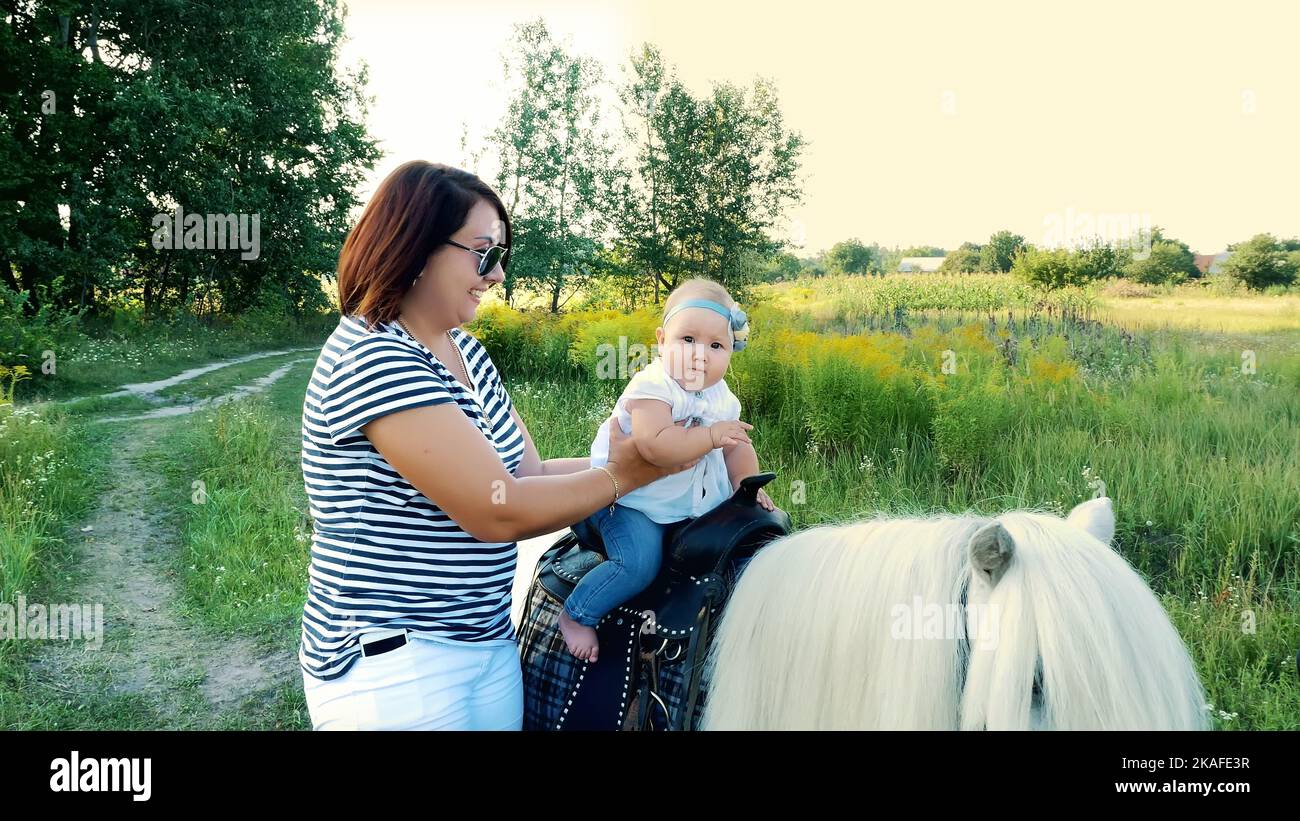 Eine Frau mit einem Baby geht um das Feld, ein Baby sitzt auf einem Pony, Mama hält das Baby. Fröhlicher, glücklicher Familienurlaub. Im Freien, im Sommer, in der Nähe des Waldes. Hochwertige Fotos Stockfoto