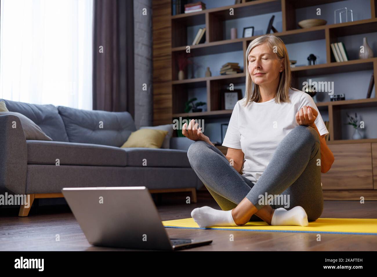 Grauhaarige, glückliche, reife Frau, die online mit dem Laptop Gymnastik und Yoga zu Hause macht. Aktiver und gesunder Lebensstil für Rentner. Fitnesskurse Stockfoto
