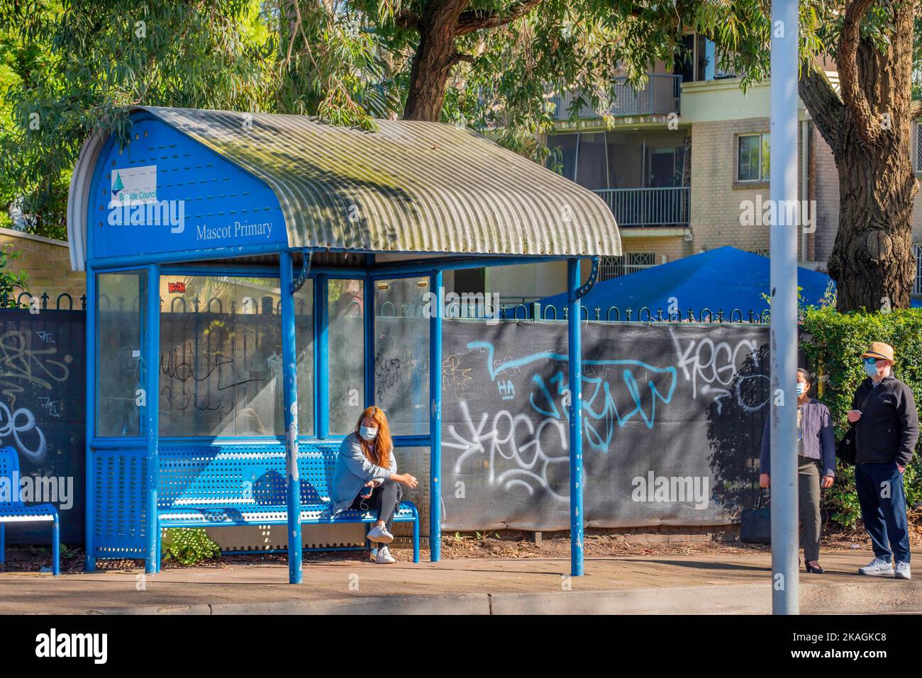 Eine Frau sitzt und zwei weitere Personen stehen und warten an einer Bushaltestelle an der Botany Road in Mascot, Sydney, Australien, auf einen Bus Stockfoto