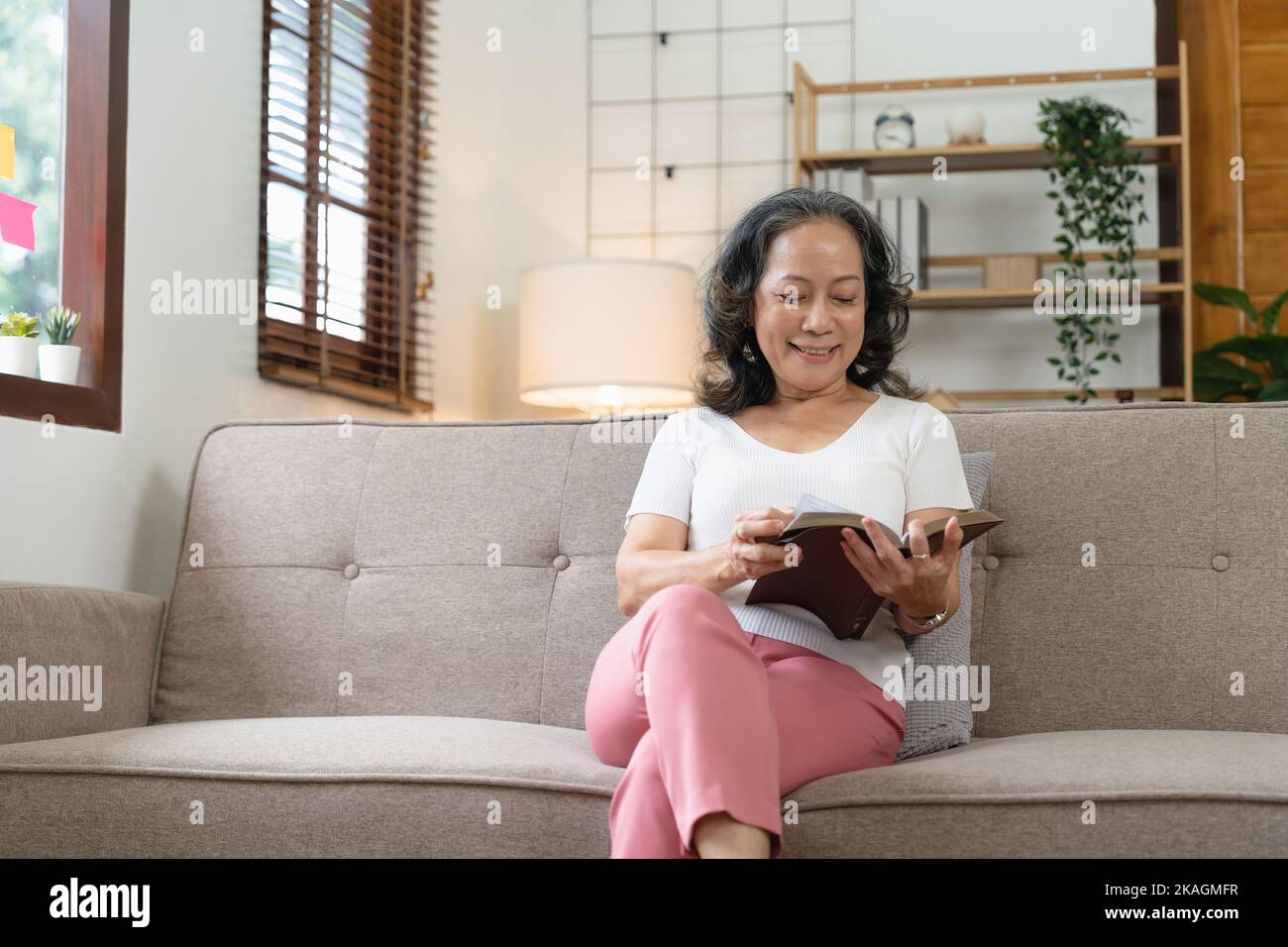 Lächelnde attraktive Rentnerin liest Buch, während sie zu Hause auf dem Sofa sitzt. Stockfoto