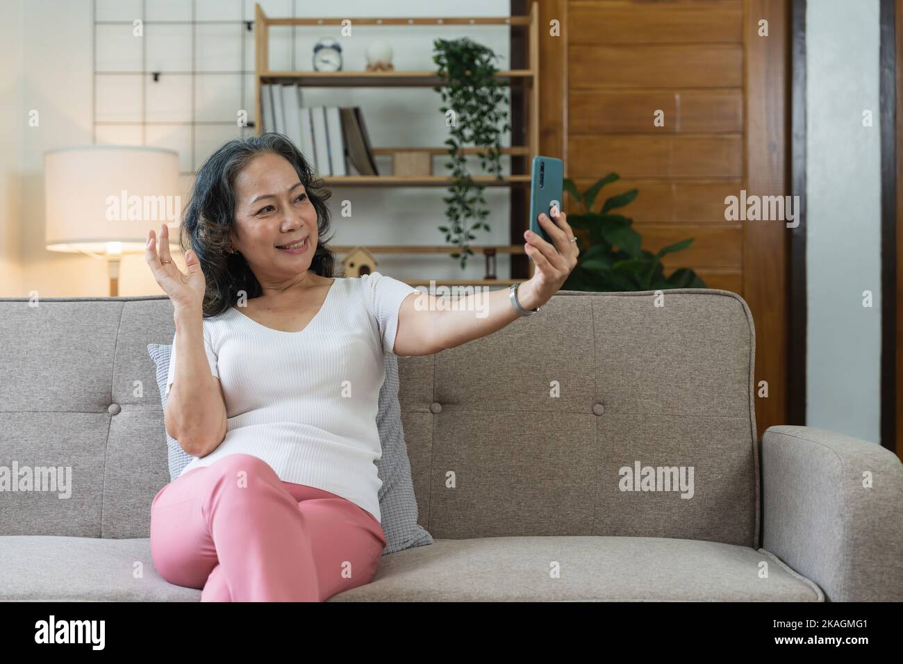 Asiatische ältere Frau, die Selfie auf dem Handy nimmt, während sie auf dem Sofa sitzt und lächelt Stockfoto