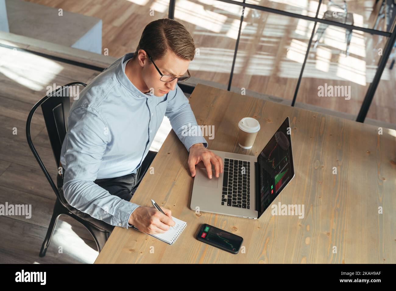 Draufsicht auf einen gutaussehenden Geschäftsmann, der online arbeitet und am Tisch im modernen Büro sitzt und Informationen auf dem Notizblock notiert, nachdem er die Preisprognosstabelle auf seinem Laptop und seiner Telefon-App überprüft hat Stockfoto