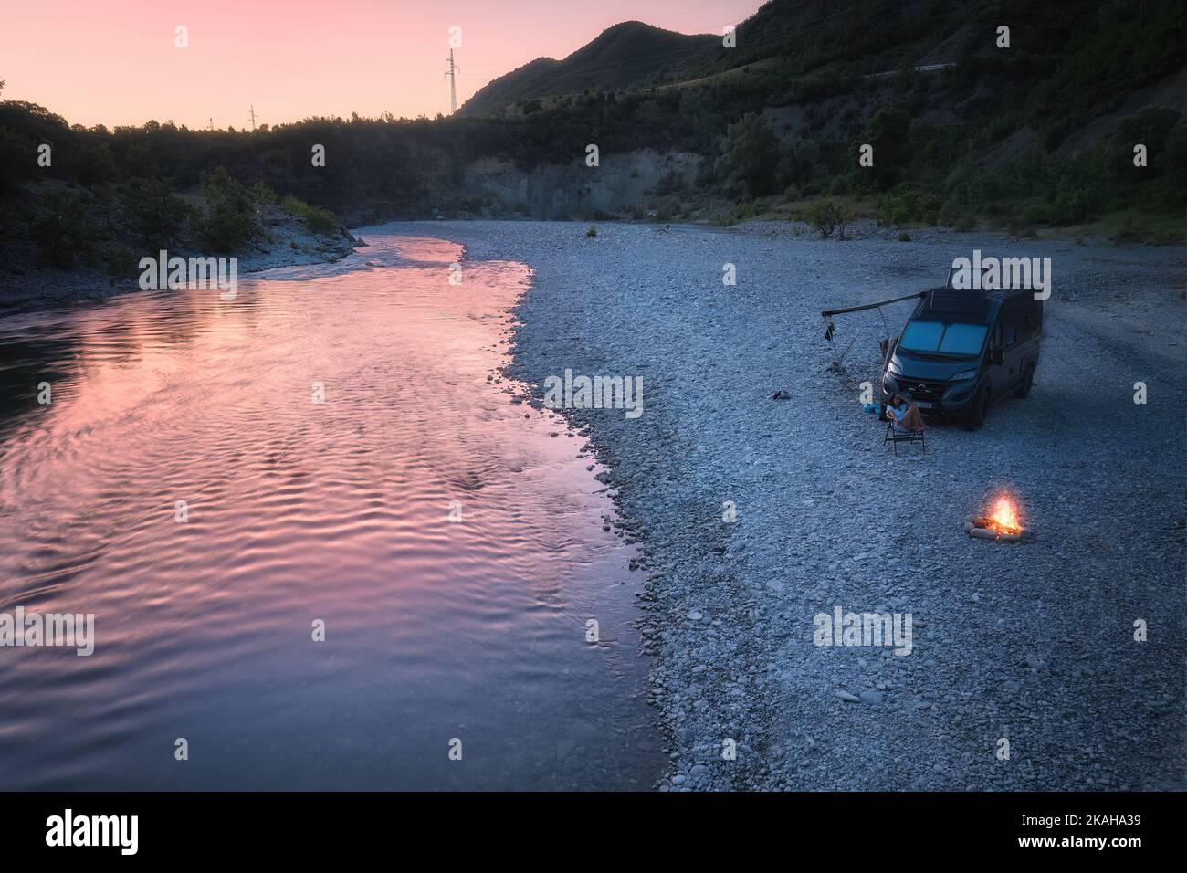 Luftaufnahme des Wohnwagens am wilden Flussstrand vor rosa Sonnenuntergang. Lagerfeuer, wildes Campen in der Natur. Outdoor Nomaden Lifestyle. Stockfoto