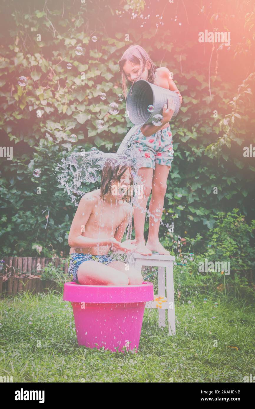 Junge, der auf einem Hocker steht und einen Eimer kaltes Wasser über einen anderen Jungen im Garten gießt Stockfoto