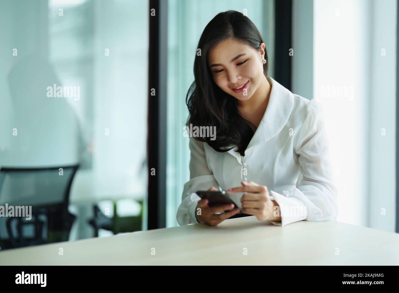 Porträt einer jungen asiatischen Frau, die mit ihrem Telefon an einem Schreibtisch sitzt Stockfoto