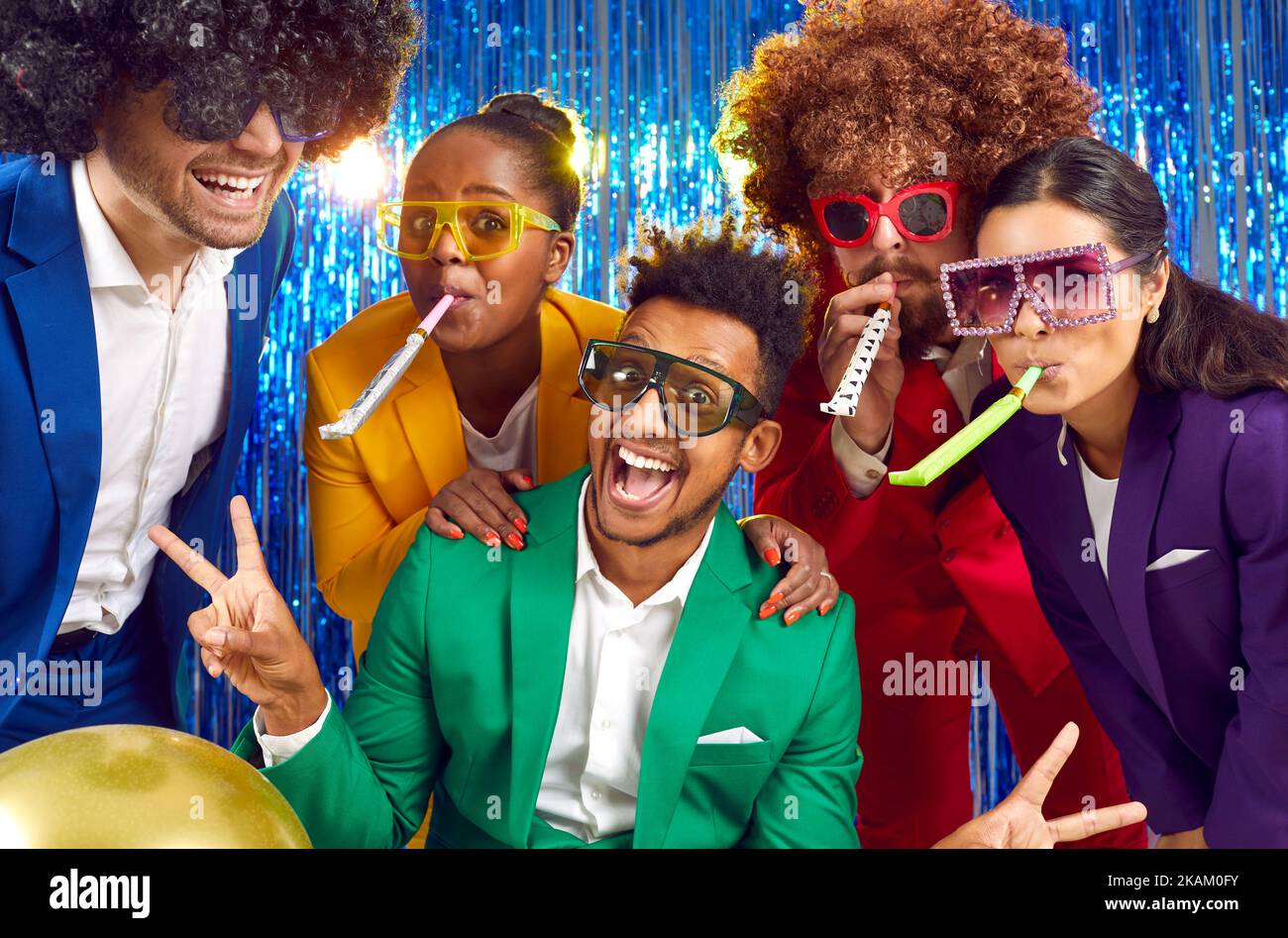 Fröhliche Menschen in bunten Kostümen, witzigen Disco-Brillen und Perücken, die gemeinsam Party-Hörner blasen Stockfoto
