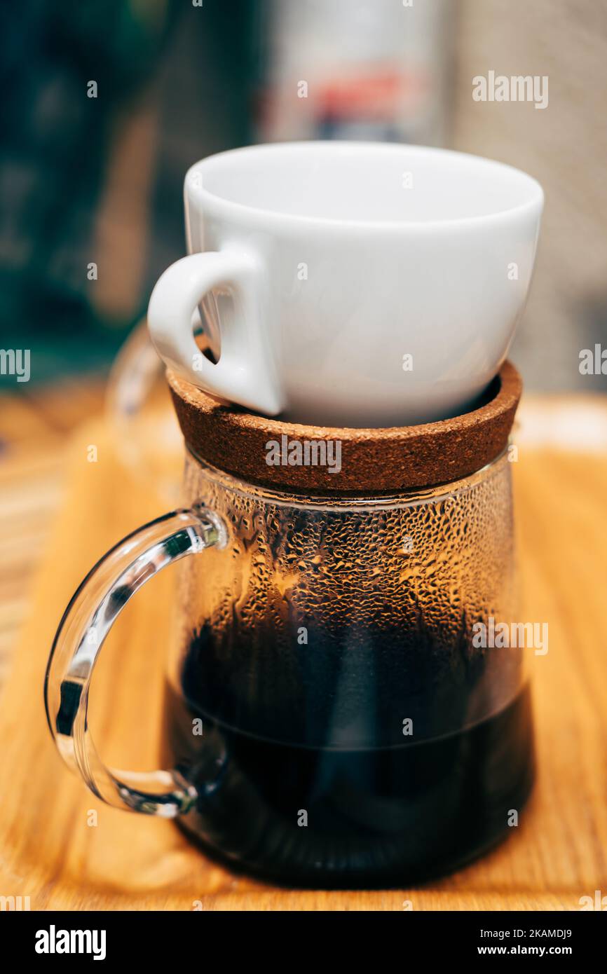 Vertikale Aufnahme von Glas-Kaffee-Server mit frischem leckeren Kaffee und einer weißen Tasse darüber auf einem Holztisch. Stockfoto