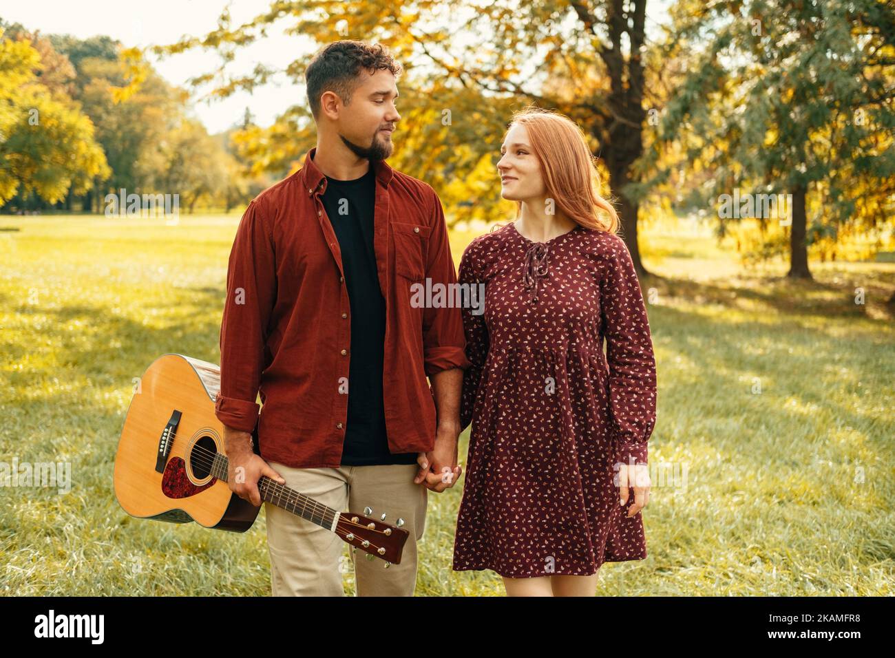 Ein junges, glückliches Paar geht mit einer akustischen Gitarre in roten Kleidern in einem sonnigen grünen Park spazieren. Stockfoto