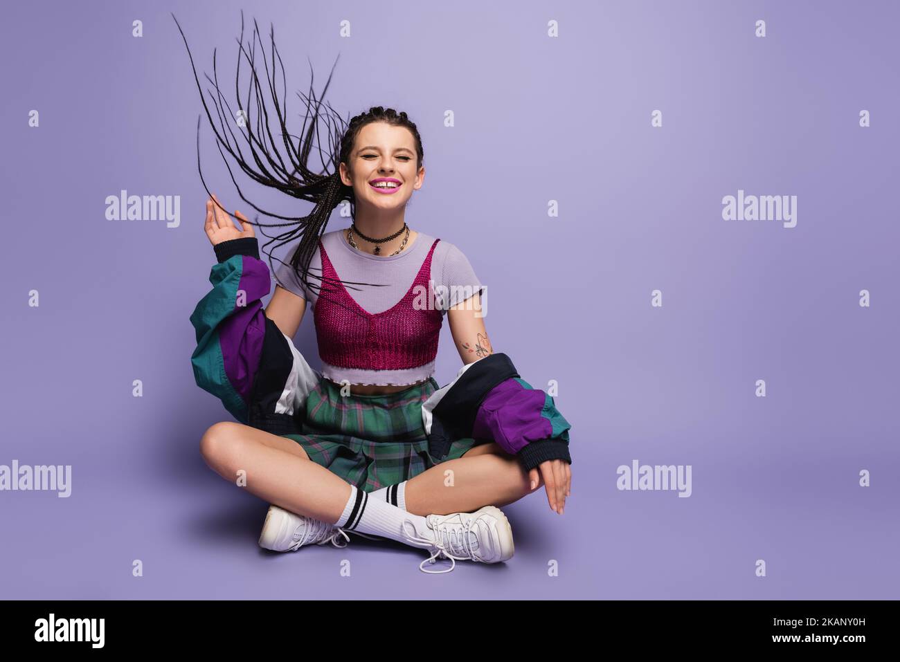 Lächelnde Frau im Vintage-Outfit winkende Hand, während sie auf Wind mit gekreuzten Beinen auf purpurem Hintergrund sitzt, Stockbild Stockfoto