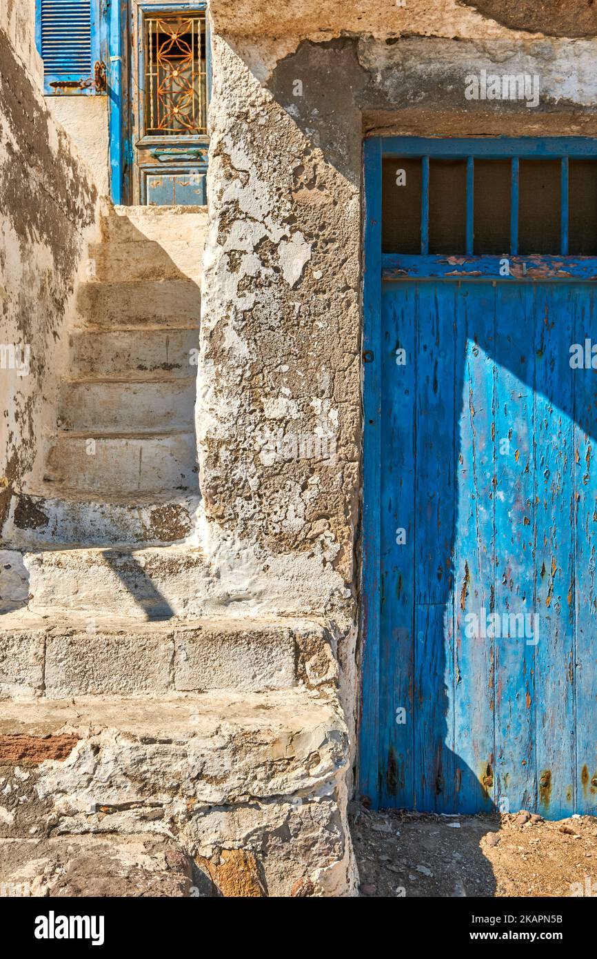 Verblasste und verwitterte weiße Wände und Treppen, blau gestrichene Tür, Griechenland. Traditionelle Architektur, Details, authentische Fassade, sonniger Tag, Sonnenschein, Schatten Stockfoto