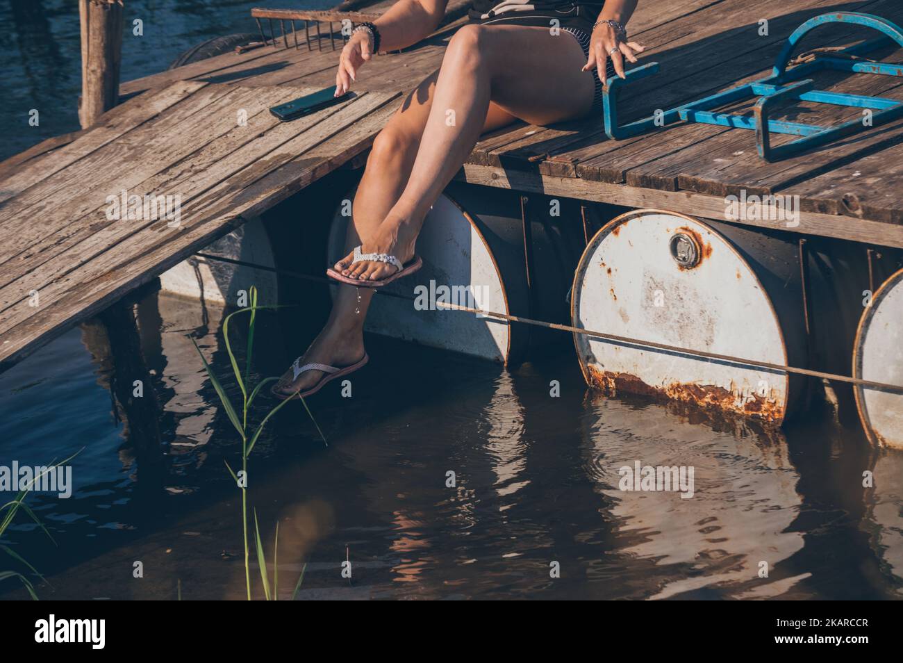 Gebräunte Beine einer Frau, die auf einer Brücke sitzt und die Füße im Flusswasser wascht Stockfoto