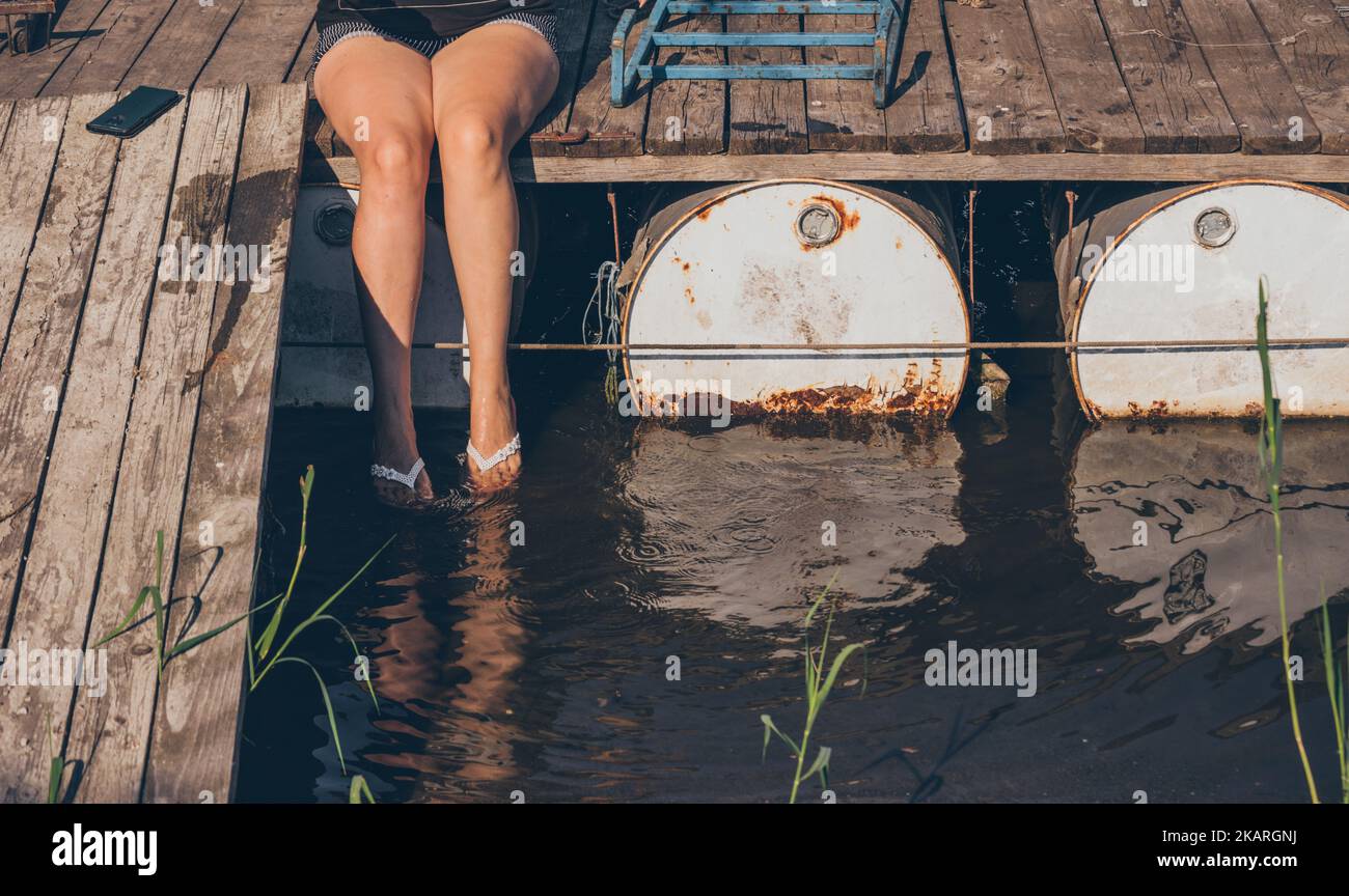 Beine einer Frau, die auf einer alten Holzbrücke über dunklem Wasser sitzt Stockfoto
