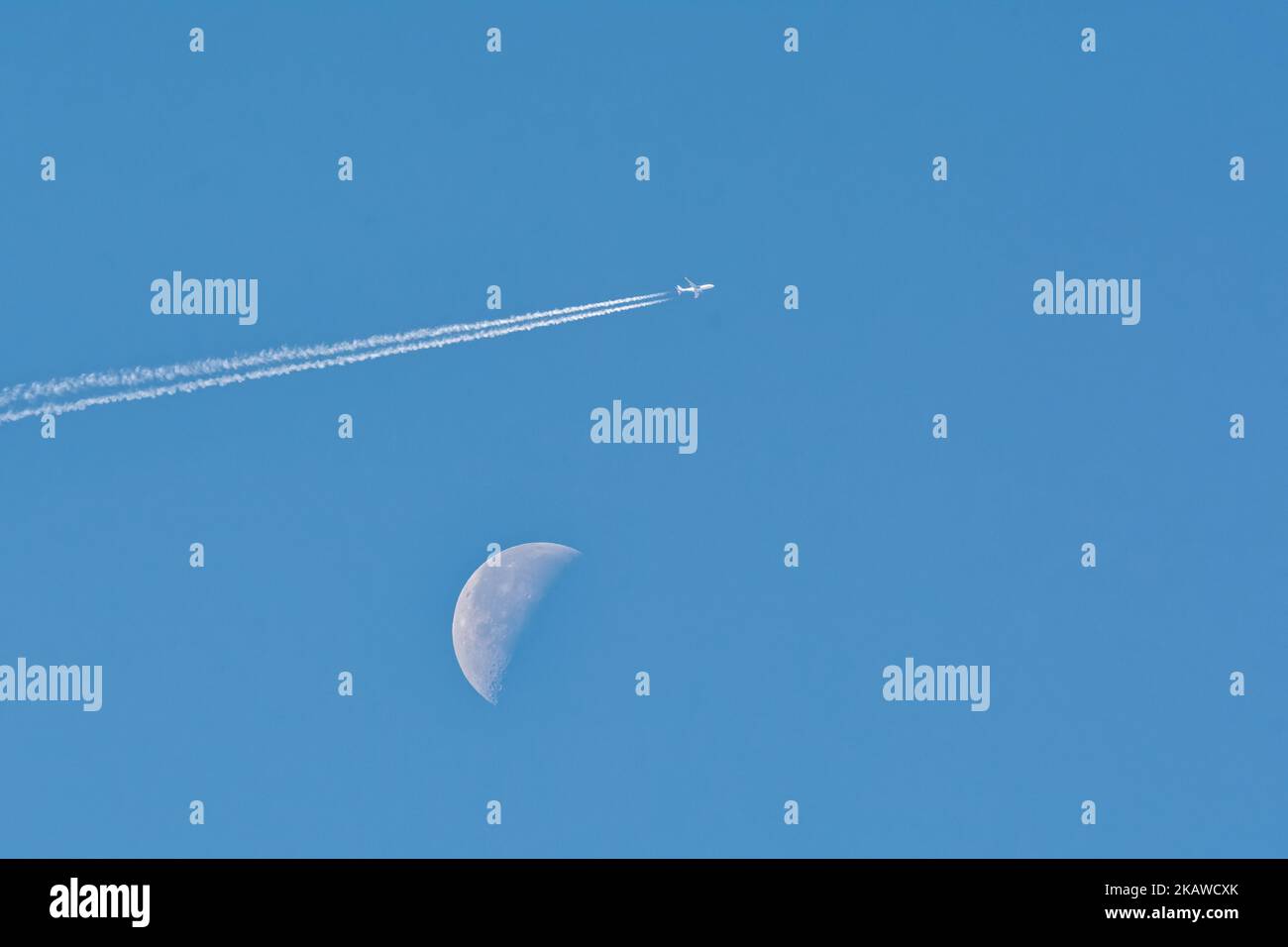 Hochfliegender Passagierjet-Liner mit nachliegendem Kondensstreifen, der einen Halbmond vor einem klaren, blauen Himmel passiert Stockfoto