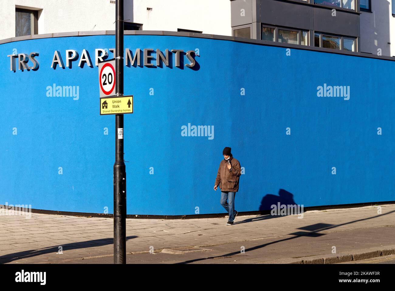 Eine männliche Figur, die an einem kräftigen blau bemalten Horten in starkem Sonnenlicht vorbeigeht und ein einfaches abstraktes Muster in einer städtischen Straße in Southall London schafft Stockfoto