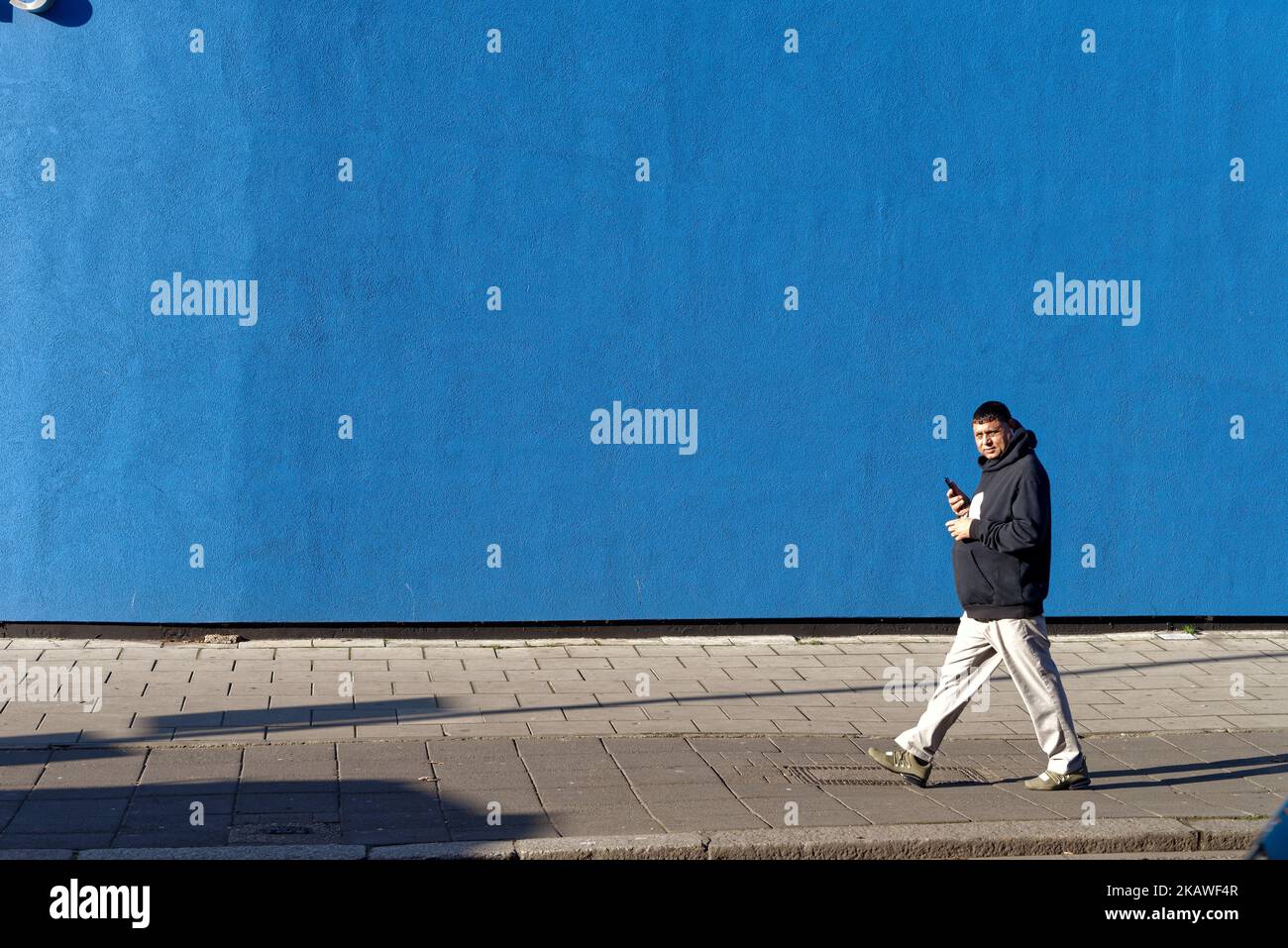 Eine männliche Figur, die an einem kräftigen blau bemalten Horten in starkem Sonnenlicht vorbeigeht und ein einfaches abstraktes Muster in einer städtischen Straße in Southall London schafft Stockfoto