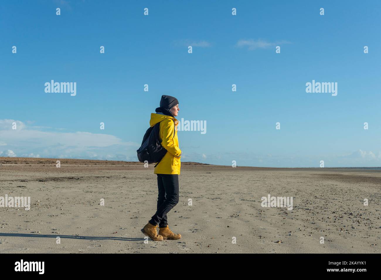 Frau mit gelbem Mantel und Rucksack, die am Strand entlang läuft, sonniger Herbsttag. Stockfoto