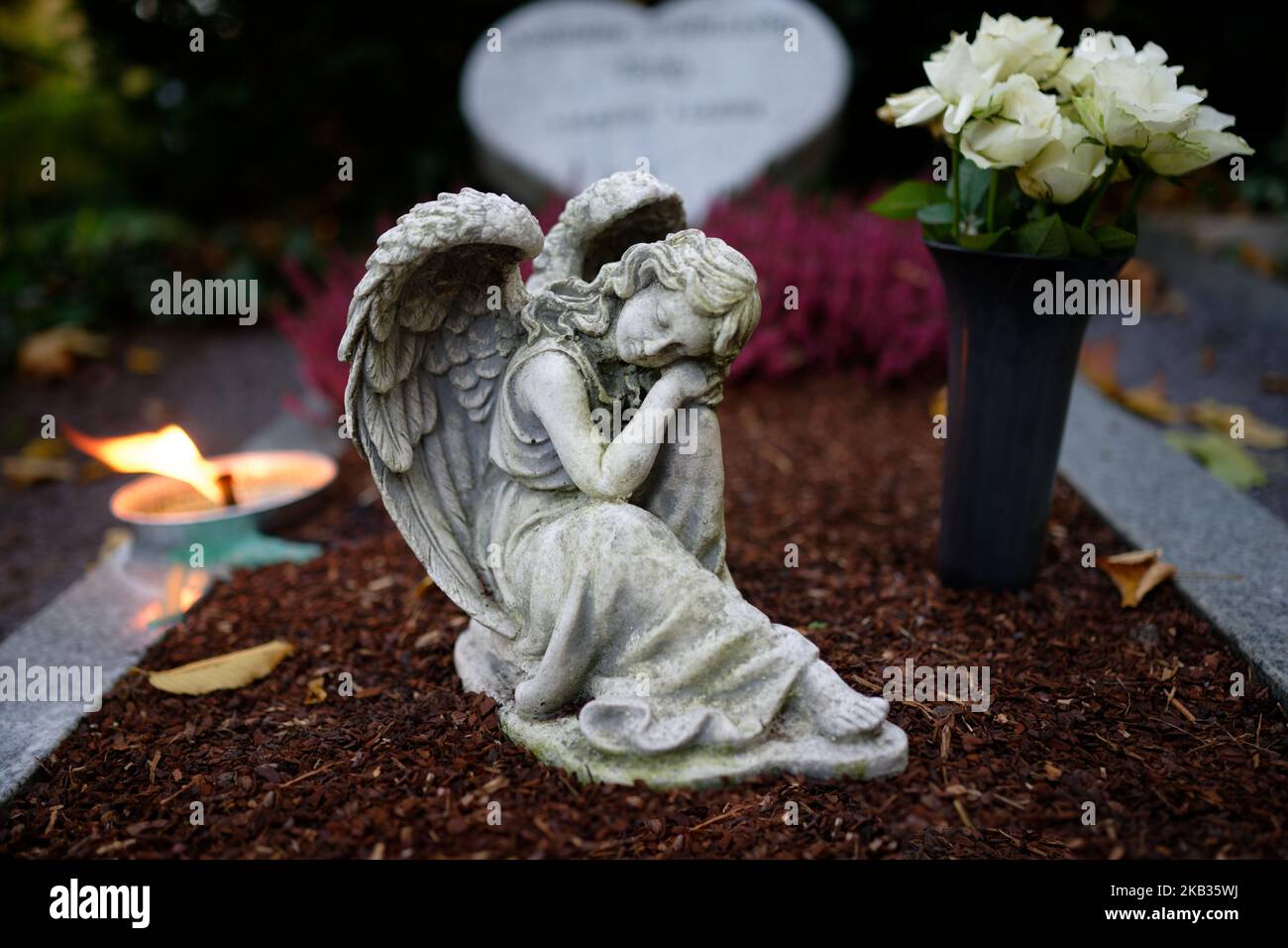 Kleine Engelsfigur mit Flügeln auf einem Grab mit Blumen und brennender Kerze in der Dämmerung Stockfoto