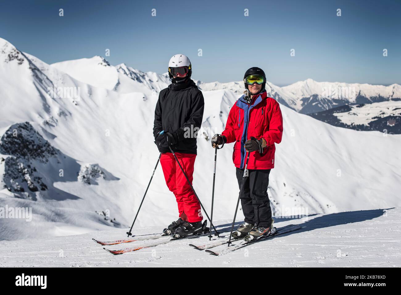Zwei Skifahrer eine Frau ein Mann hält am Rand des Berges das Foto, hohe Berge der Alpen im Hintergrund auf einem sonnigen Tag Helme und Brillen auf roten und schwarzen Outfits Stockfoto