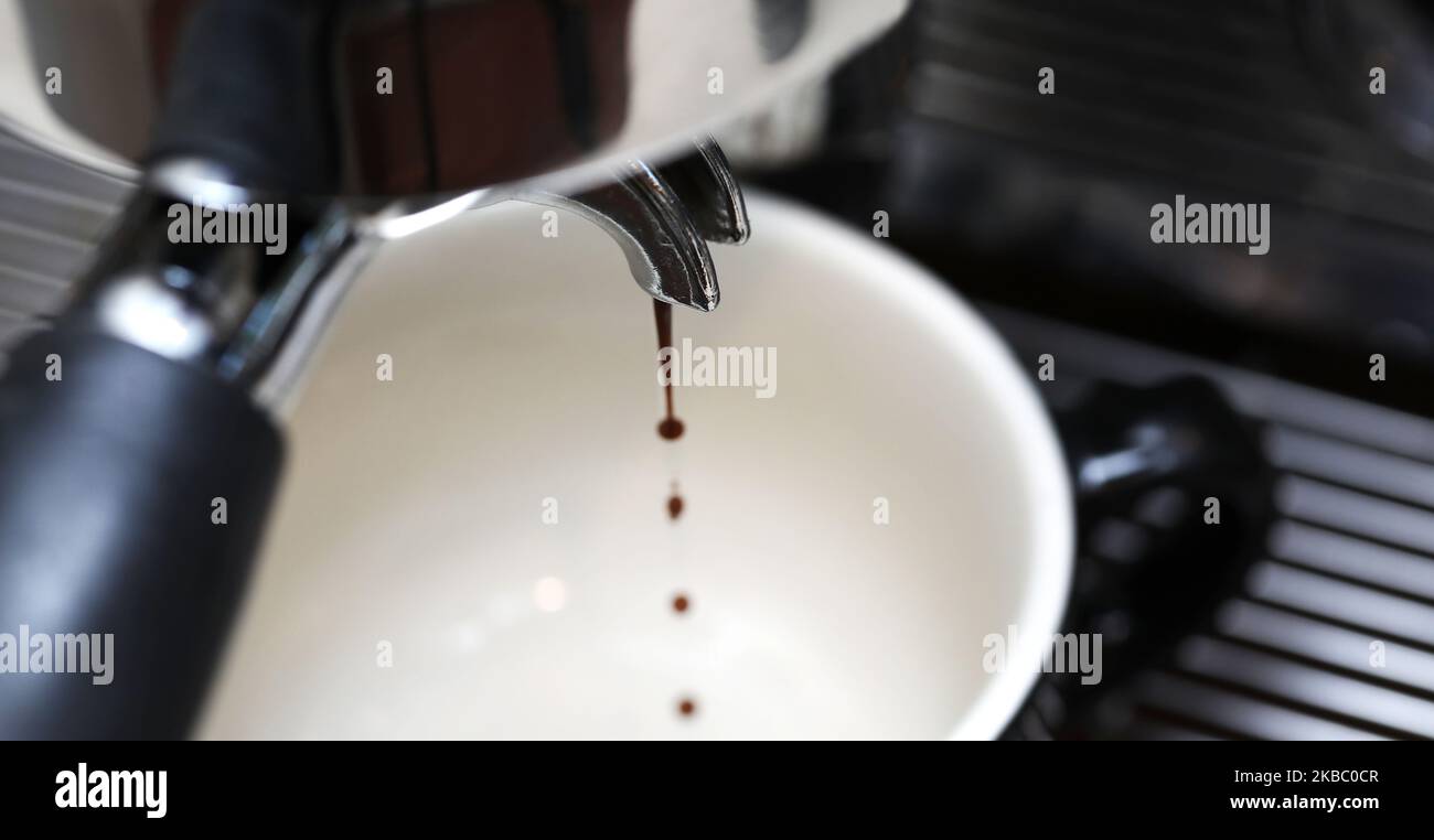 Eine Nahaufnahme der Filterspitze, die die ersten Tropfen aus dem perfekten Espresso-Kaffee in eine Tasse mit schwarzem Griff gießt. Stockfoto